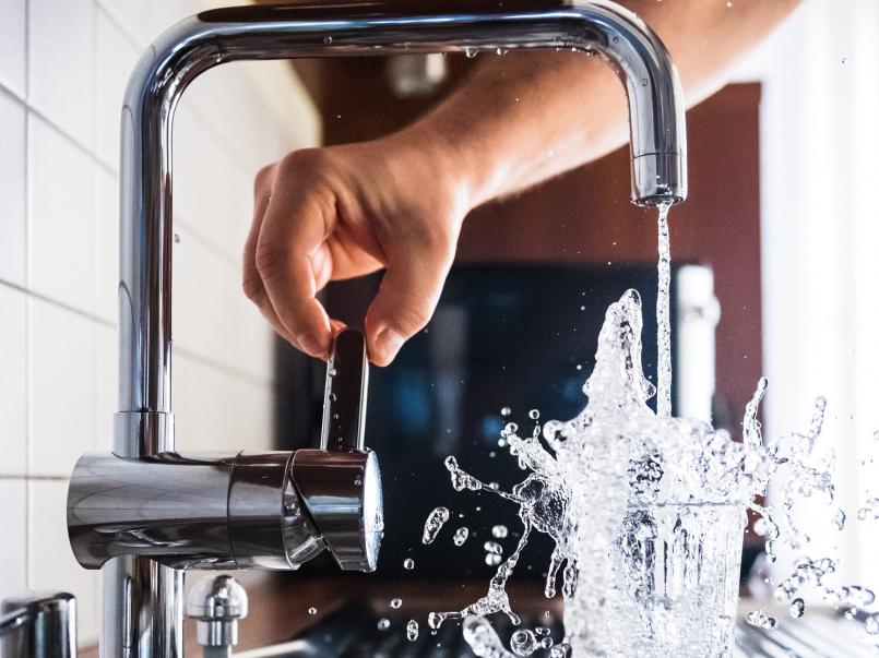 A arm and hand turning on a tap which overspills into a glass of water