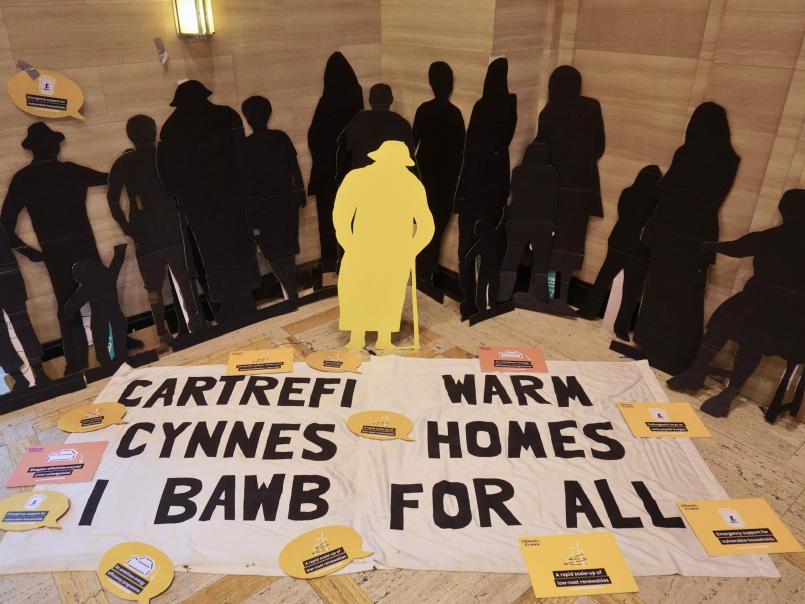 Black silhouettes behind a sign that says 'Warm Homes for all' in English and Welsh/Silwetau du tu ôl i arwydd sy'n dweud 'Cartrefi Cynnes i Bawb' yn Gymraeg a Saesneg