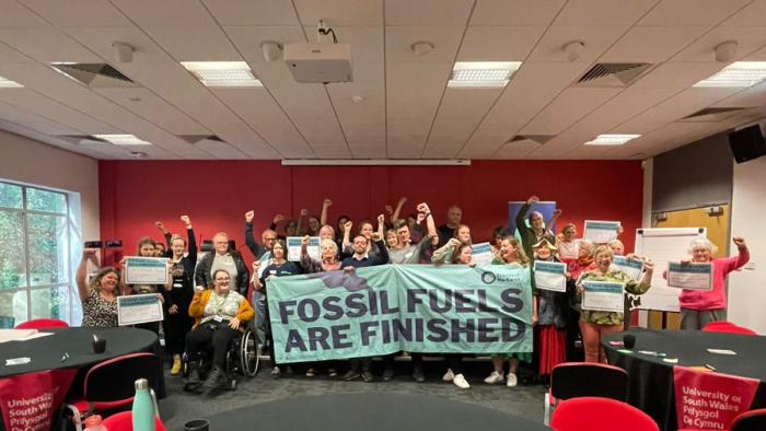 Crowd of people inside a conference venue standing behind a banner, 'Fossil Fuels are Finished' with their arms in the air